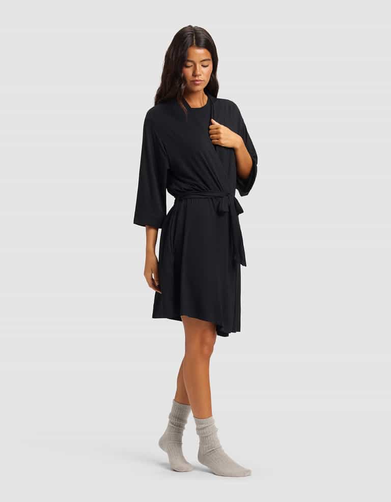 A woman with long dark hair stands against a plain background wearing the Cozy Earth Women's Bamboo Stretch-Knit Kimono Robe in black, featuring three-quarter sleeves and a tie waist, paired with light gray socks. She has a relaxed posture.|Color:Black