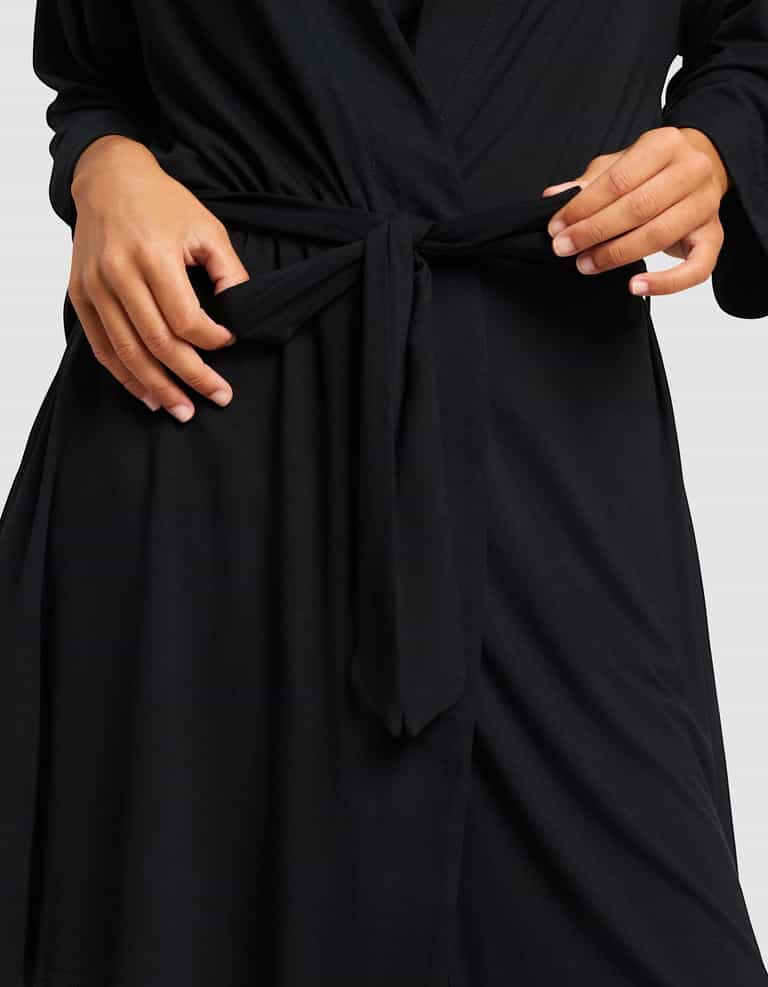 A person adjusts the waist tie of a Cozy Earth Women's Bamboo Stretch-Knit Kimono Robe, showing only their torso and hands against a plain background.|Color:Black