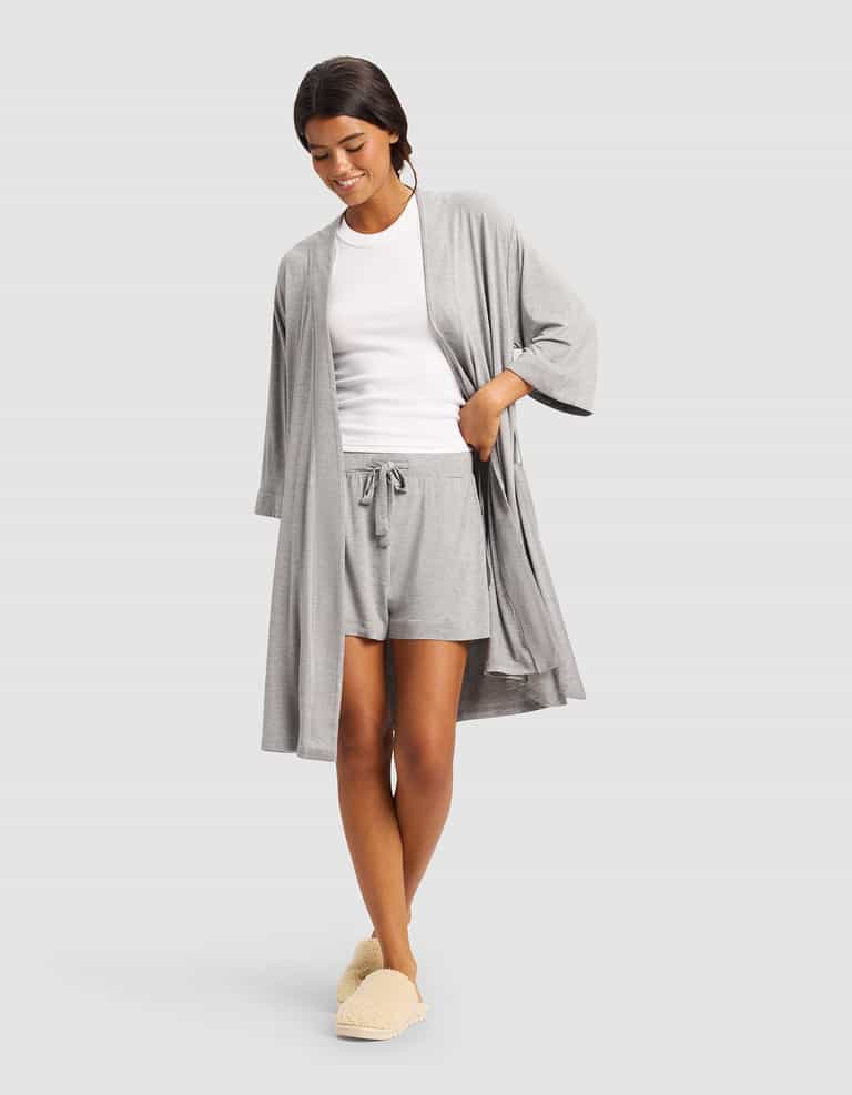 A woman wearing Cozy Earth’s Women's Bamboo Stretch-Knit Kimono Robe in grey, paired with a white T-shirt, matching grey shorts, and cream slippers, smiles with one hand in her pocket against a plain light gray background.|Color:Grey