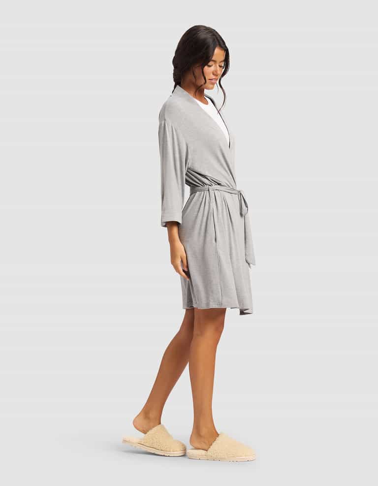 A woman stands sideways against a plain light background, relaxed in Cozy Earth’s Women's Bamboo Stretch-Knit Kimono Robe in light gray with a waist tie, paired with beige slippers. Her loose waves add to the comfortable vibe.|Color:Grey