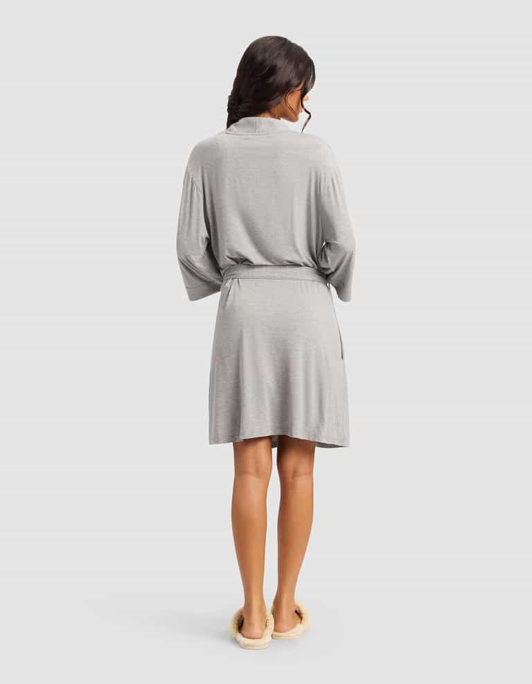 A woman with long dark hair, seen from the back, wears a Cozy Earth Women's Bamboo Stretch-Knit Kimono Robe in light gray with a belt and cream slippers, standing against a plain light background.|Color:Grey