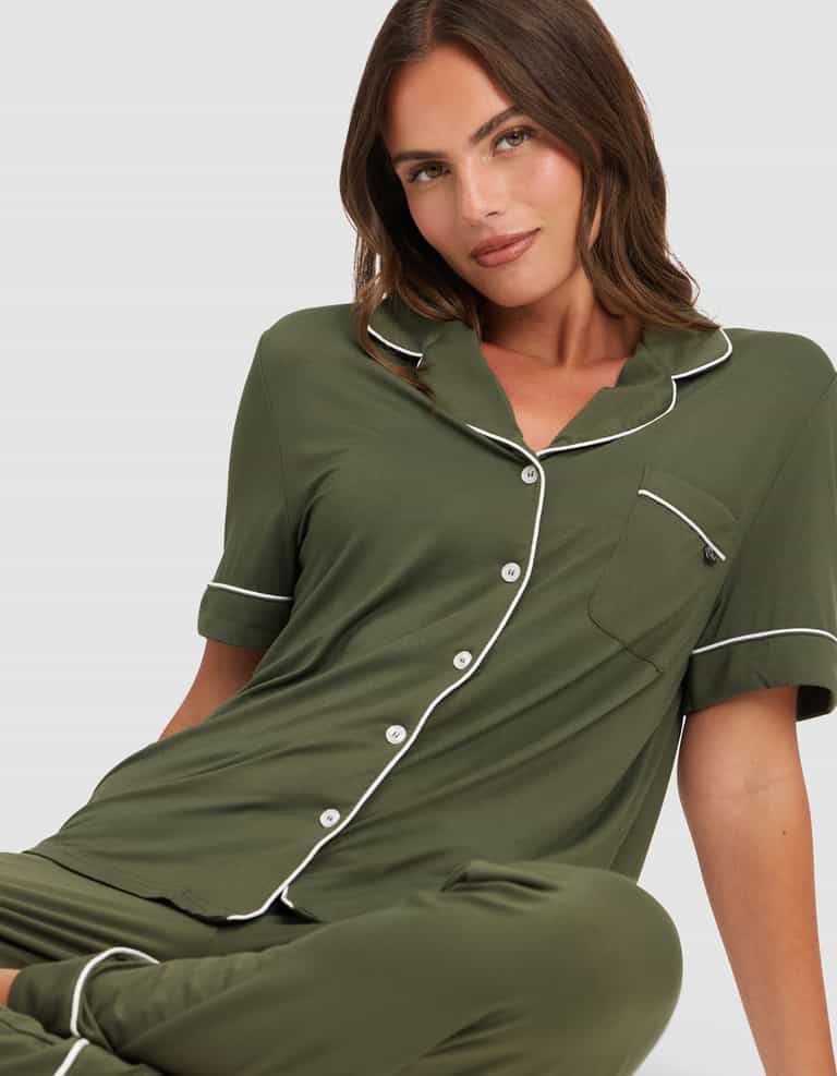 A woman with brown hair wears the HIDE Women's Bamboo Stretch-Knit Short Sleeve Pajama Top in olive green with white piping and buttons. She sits on the floor, facing the camera with a slight smile against a plain light background. |Color:Olive