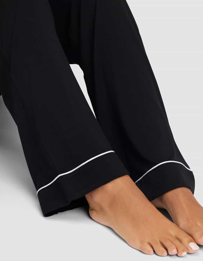 A close-up of bare feet and lower legs in Cozy Earth’s Women's Bamboo Stretch-Knit Long Sleeve Pajama Set, featuring loose black pants with white piping at the hem, set against a plain light gray background. |Color:Black