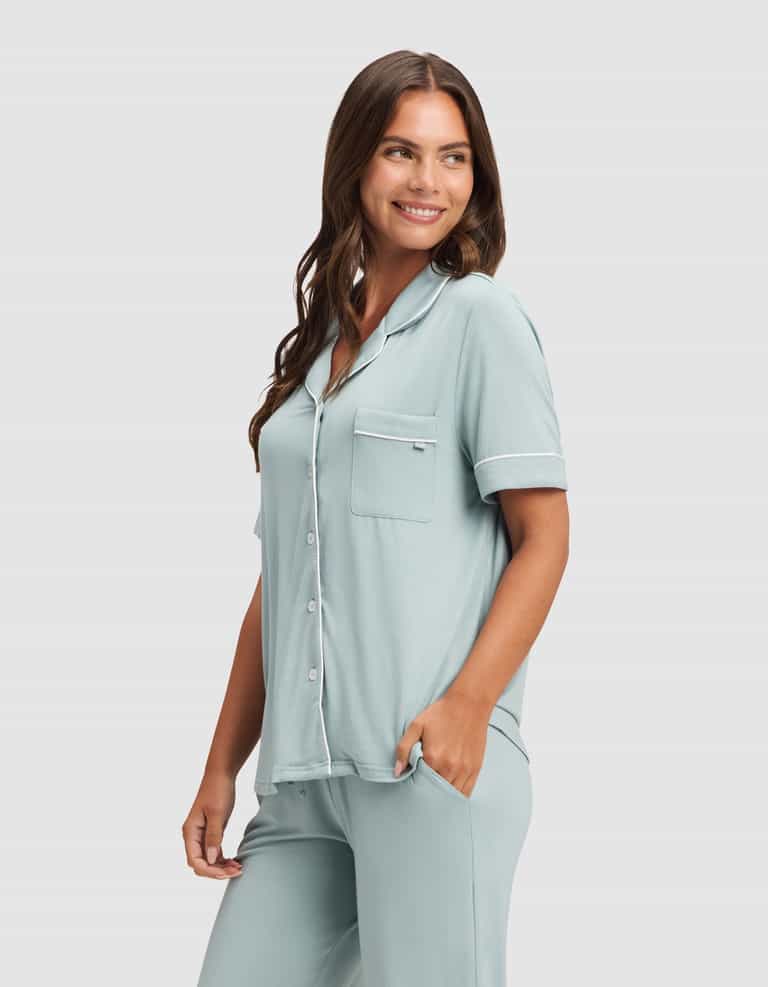 A woman smiles with one hand in her pocket, wearing the Cozy Earth Women's Bamboo Stretch-Knit Short Sleeve & Pant Pajama Set in light blue, featuring a button-up top and matching pants against a plain light background. |Color:Brook