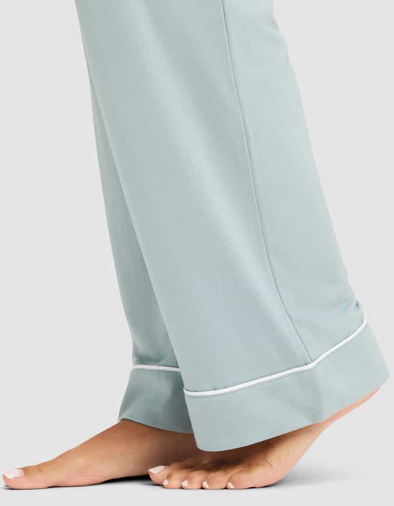 A person stands barefoot on a light background wearing Cozy Earth's Women's Bamboo Stretch-Knit Long Sleeve Pajama Set in light blue with white piping detail near the cuffs. |Color:Brook