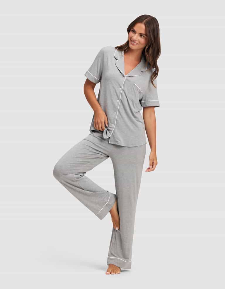 A woman models the Cozy Earth Women's Bamboo Stretch-Knit Short Sleeve & Pant Pajama Set in light gray with white trim, standing barefoot, smiling, and lifting one leg as she holds the hem of her top. |Color:Grey