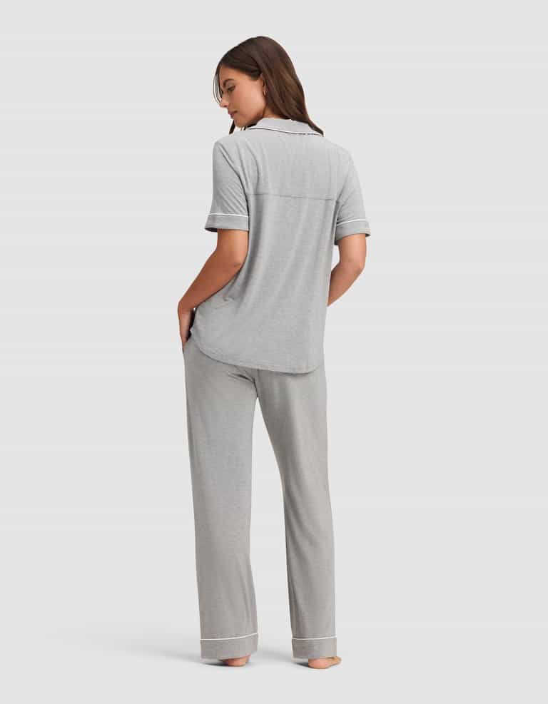A woman stands barefoot, facing away, wearing the Cozy Earth Women's Bamboo Stretch-Knit Short Sleeve & Pant Pajama Set in light gray, featuring cuffed long pants. She has one hand in her pocket and her head turned slightly to the side. |Color:Grey