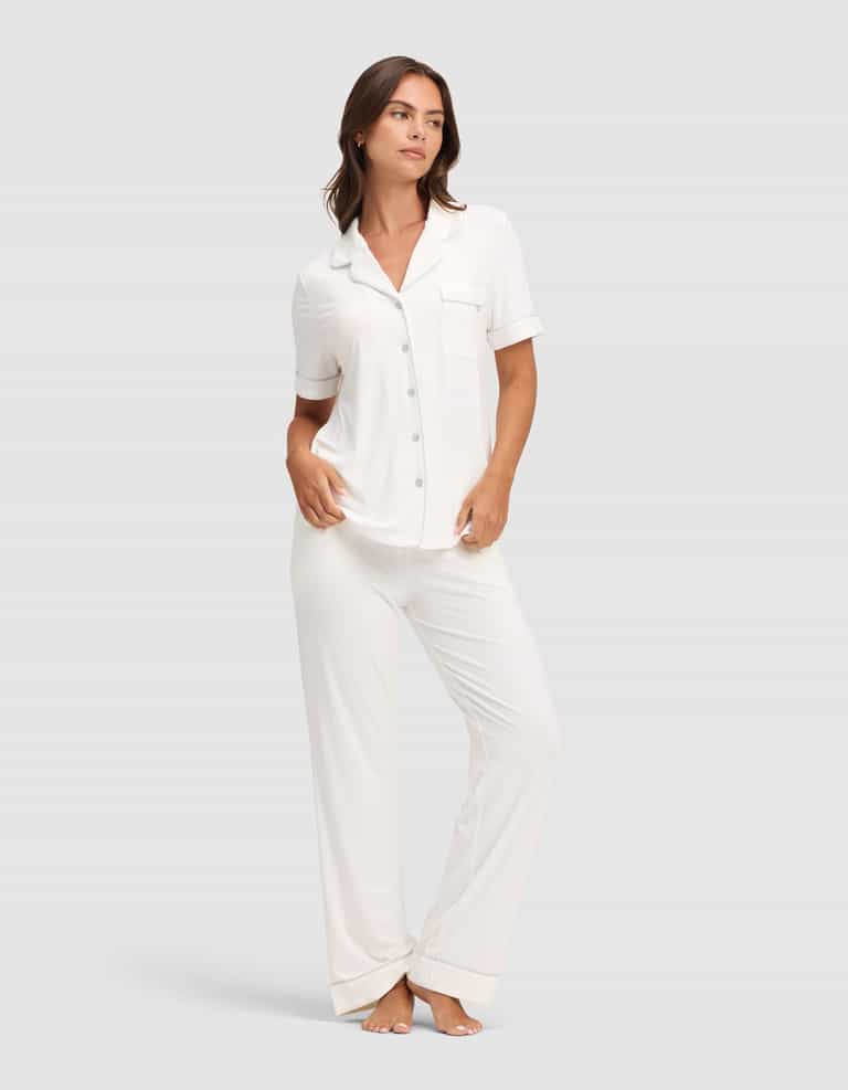 A woman wears Cozy Earth's Women's Bamboo Stretch-Knit Short Sleeve & Pant Pajama Set, featuring a white short-sleeved button-up top with chest pocket and matching long pants, standing barefoot against a plain light gray background. |Color:Ivory