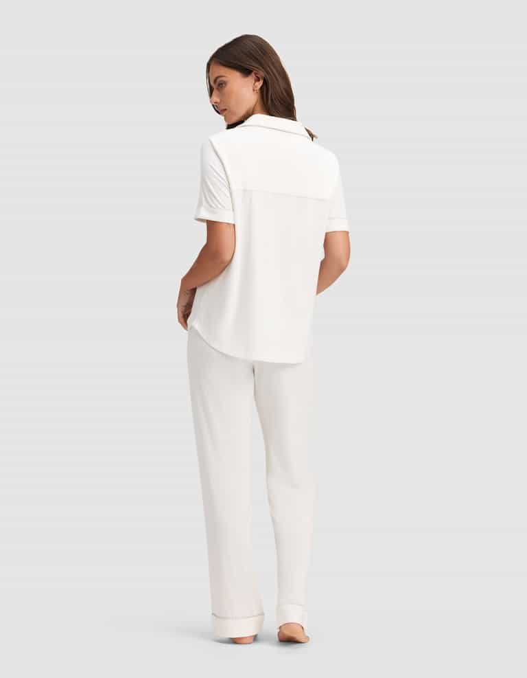A woman faces away, barefoot on a light gray background, wearing Cozy Earth's Women's Bamboo Stretch-Knit Short Sleeve & Pant Pajama Set in white. |Color:Ivory