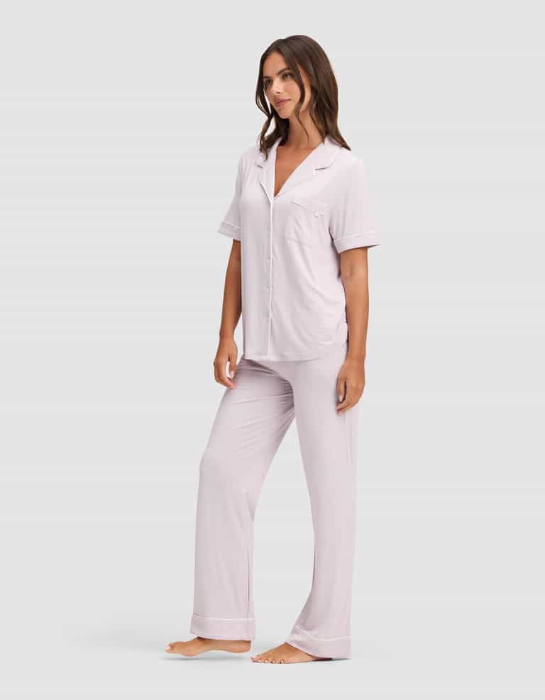 A woman stands barefoot in Cozy Earth's Women's Bamboo Stretch-Knit Short Sleeve & Pant Pajama Set, featuring a light pink button-up shirt and long pants, against a plain light gray background.|Color:Lilac
