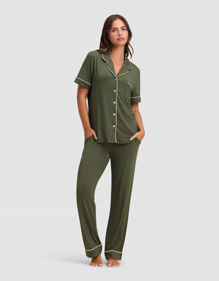 A woman stands barefoot in the Cozy Earth Women's Bamboo Stretch-Knit Short Sleeve & Pant Pajama Set in olive green with white piping, featuring a button-up top and long pants, her hands in her pockets against a plain light background.|Color:Olive