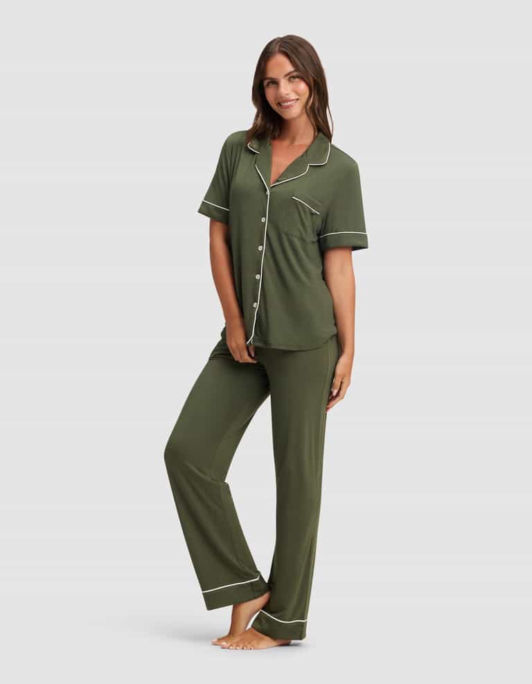 A barefoot woman smiles, wearing Cozy Earth's Women's Bamboo Stretch-Knit Short Sleeve & Pant Pajama Set in olive green with white piping, featuring a button-up top with a chest pocket and matching long pants.|Color:Olive