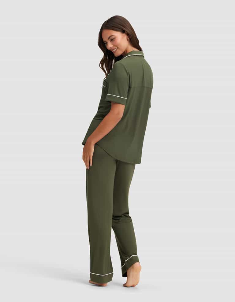 A woman stands barefoot, smiling over her shoulder, wearing the Cozy Earth Women's Bamboo Stretch-Knit Short Sleeve & Pant Pajama Set in olive green with white piping. The background is plain light gray.|Color:Olive