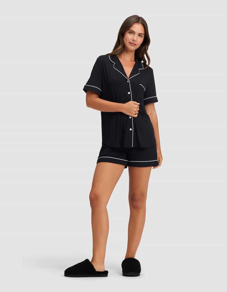 A woman wears the Cozy Earth Women's Bamboo Stretch-Knit Short Sleeve Pajama Set in black with white piping, featuring a button-up top and matching shorts, paired with black slippers against a plain light background. |Color:Black