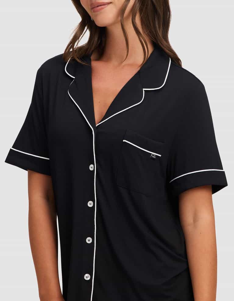A woman wears the Cozy Earth Women's Bamboo Stretch-Knit Short Sleeve Pajama Set in black, featuring white piping, front buttons, and a chest pocket, standing against a plain light background. |Color:Black