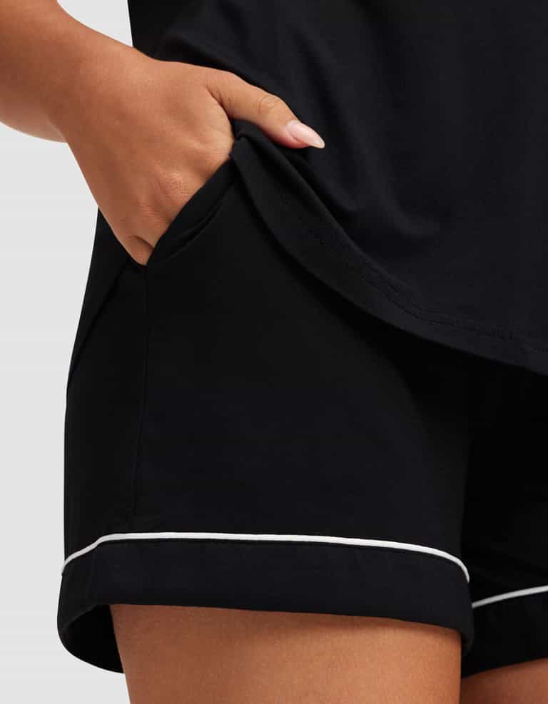 A person’s hand rests in the pocket of Cozy Earth’s Women’s Bamboo Stretch-Knit Short Sleeve Pajama Set—black shorts with white piping and a matching top. Only the lower torso, hand, and part of the outfit are visible. |Color:Black