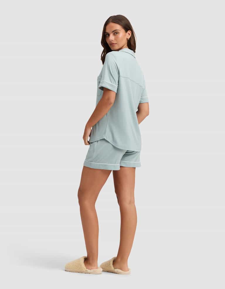 A woman, turned slightly to the side, wears the Cozy Earth Women's Bamboo Stretch-Knit Short Sleeve Pajama Set in light blue with beige slippers, posed against a plain light background. |Color:Brook