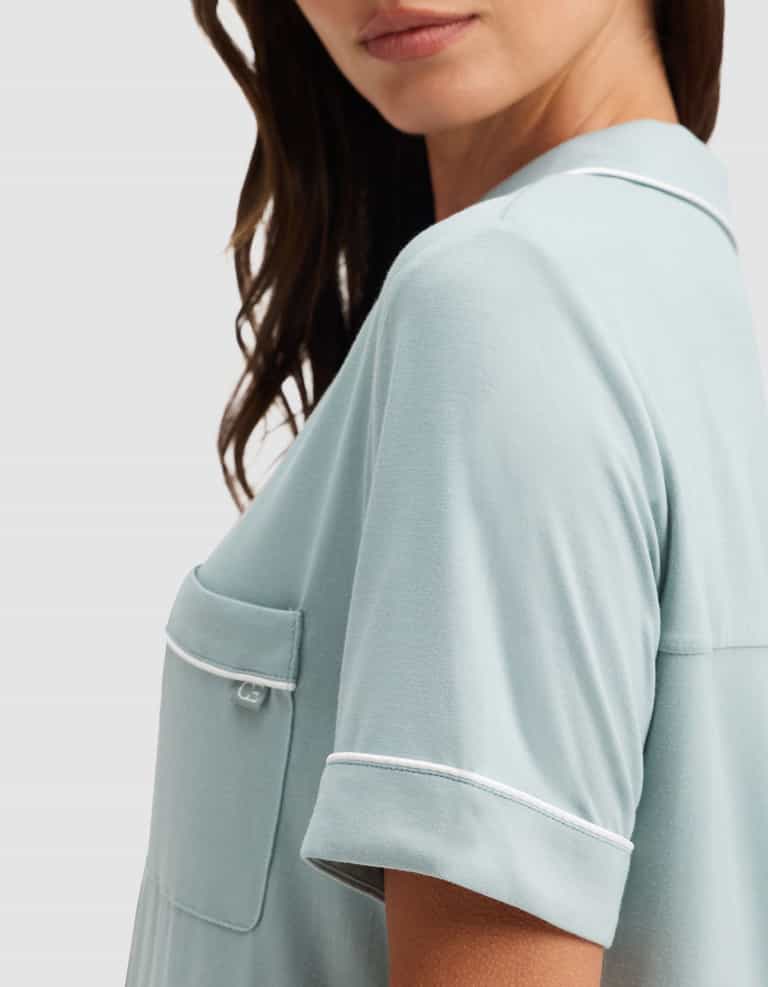 A woman with wavy brown hair models the Cozy Earth Women's Bamboo Stretch-Knit Short Sleeve Pajama Set in light blue, featuring white piping on the pocket and sleeves. The image is cropped from nose to above waist against a plain background. |Color:Brook