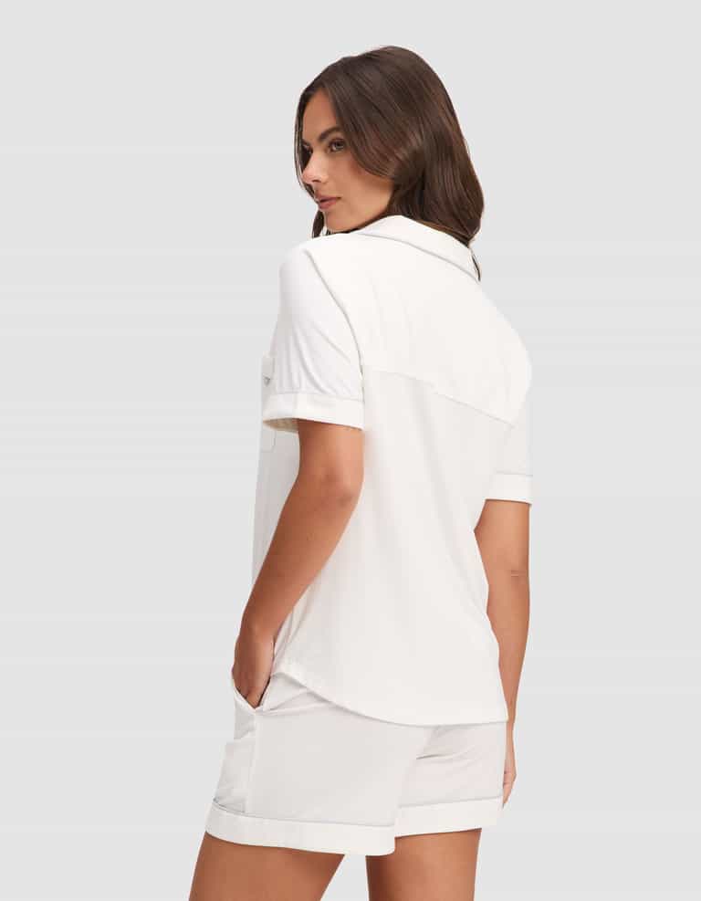 A woman with long brown hair stands sideways, looking over her shoulder, wearing the Cozy Earth Women's Bamboo Stretch-Knit Short Sleeve Pajama Set—loose-fitting white shirt and matching shorts, hands in pockets, set against a plain gray background. |Color:Ivory