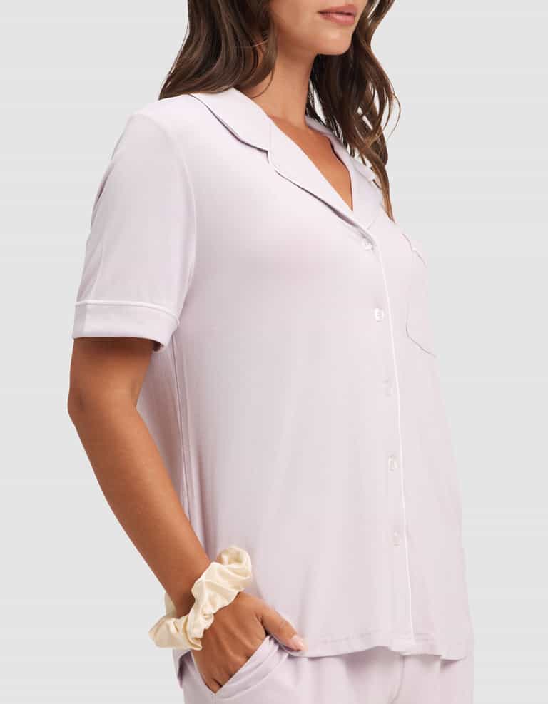 A woman wears the Cozy Earth Women's Bamboo Stretch-Knit Short Sleeve Pajama Set in light pink, featuring a chest pocket and matching bottoms. She has a beige scrunchie on her wrist and one hand in her pocket. |Color:Lilac