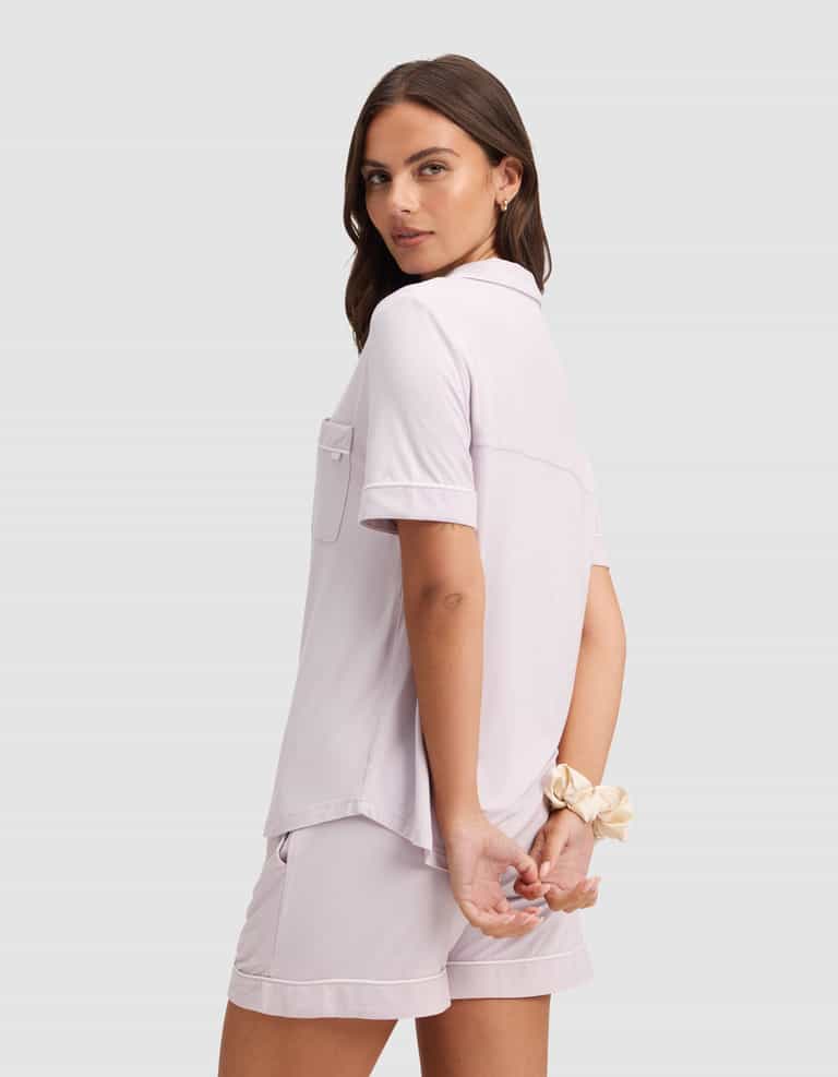 A woman models the Cozy Earth Women's Bamboo Stretch-Knit Short Sleeve Pajama Set in light lavender, standing with her back slightly turned, hands clasped behind her, and a beige scrunchie on her wrist. |Color:Lilac