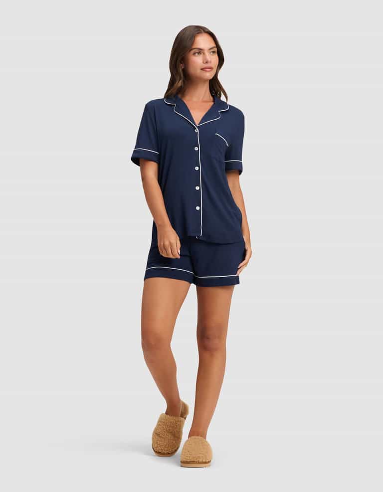 A woman wears the Cozy Earth Women's Bamboo Stretch-Knit Short Sleeve Pajama Set in navy with white piping and shorts, paired with tan fuzzy slippers, looking relaxed against a plain light background. |Color:Navy