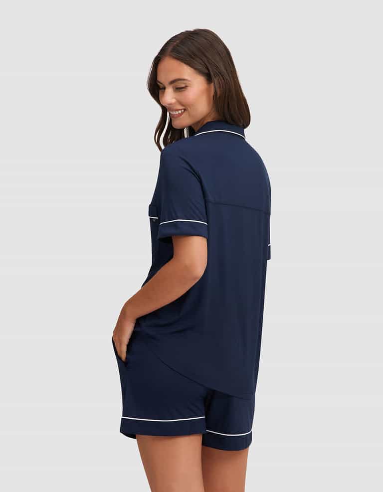 A woman with long brown hair models the Cozy Earth Women's Bamboo Stretch-Knit Short Sleeve Pajama Set in navy blue with white piping, featuring a button-up top and shorts. She smiles, glancing over her shoulder against a light gray background. |Color:Navy