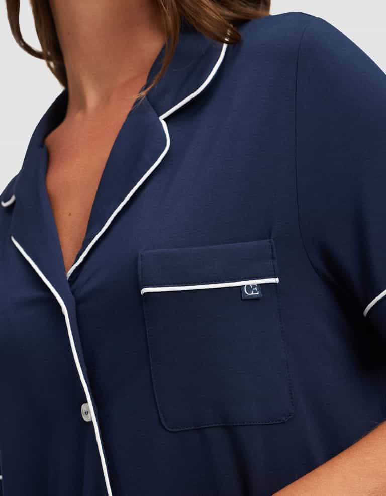 A close-up of a woman in the Cozy Earth Women's Bamboo Stretch-Knit Short Sleeve Pajama Set in navy, featuring white piping, a chest pocket with a navy and white tag, and white buttons. |Color:Navy