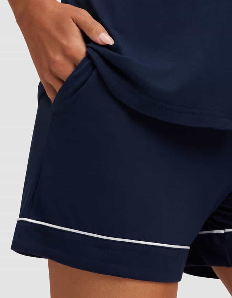 A woman models the Cozy Earth Women's Bamboo Stretch-Knit Short Sleeve Pajama Set, featuring navy blue shorts with white piping and a hand in the pocket, posed against a plain, light-colored background. |Color:Navy