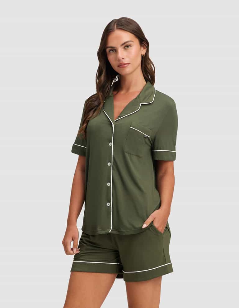 A woman wears the Cozy Earth Women's Bamboo Stretch-Knit Short Sleeve Pajama Set in dark green with white piping and buttons, standing against a plain light gray background. |Color:Olive