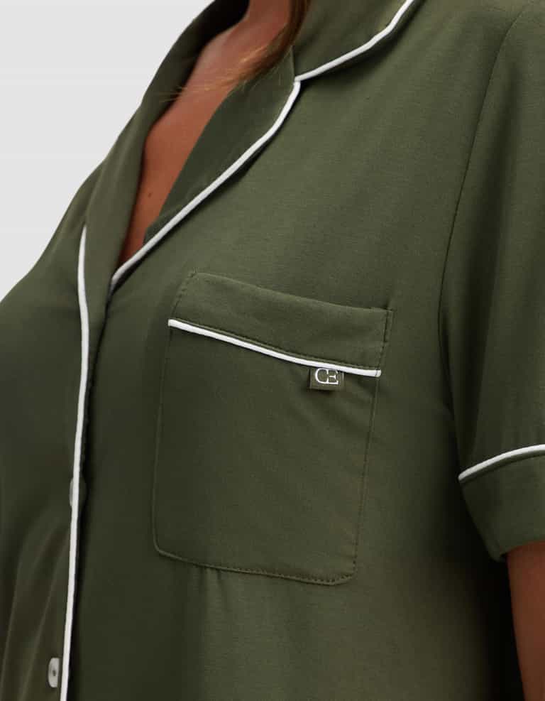 Close-up of a person wearing Cozy Earth's Women's Bamboo Stretch-Knit Short Sleeve Pajama Set in olive green, featuring white piping on the collar, pocket, and sleeves, a small chest pocket with silver tag, and white front buttons. |Color:Olive