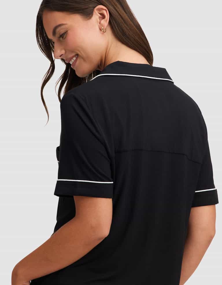 A woman with long brown hair smiles while looking down, wearing the HIDE Women's Bamboo Stretch-Knit Short Sleeve Pajama Top in black with white piping, shown from the back and side against a plain light gray background. |Color:Black