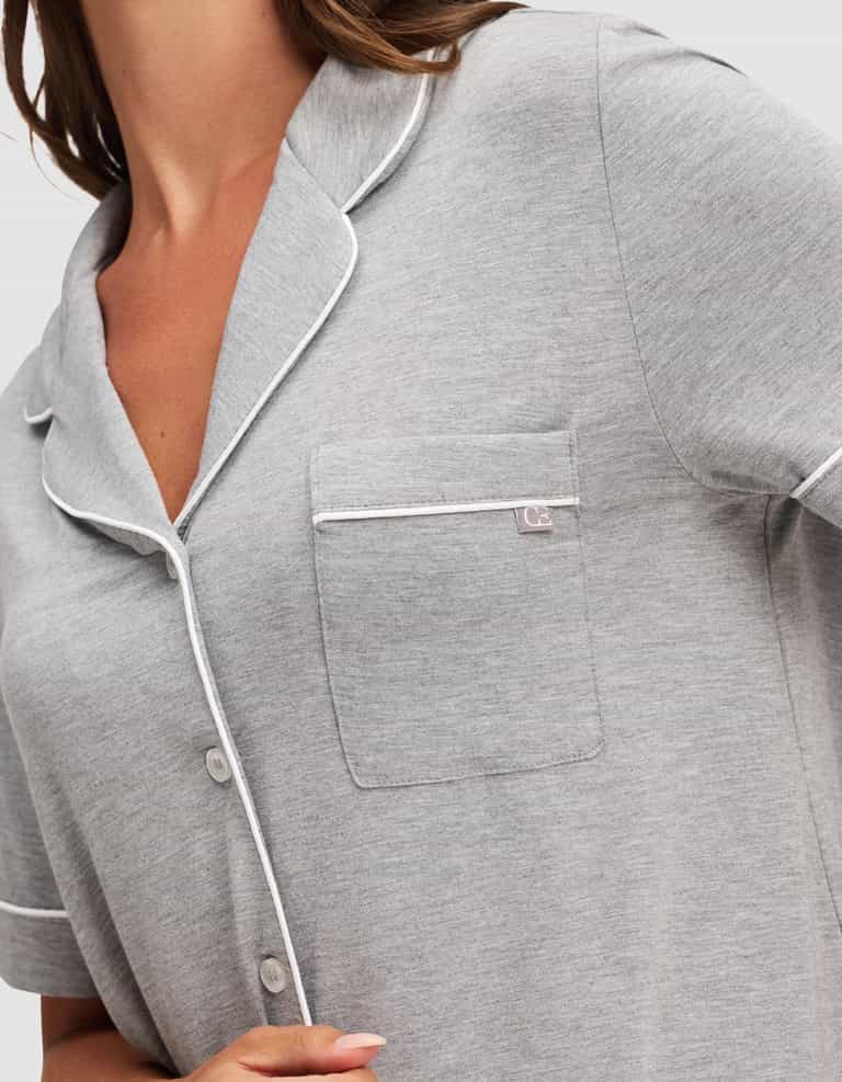 A woman wears the HIDE Women's Bamboo Stretch-Knit Short Sleeve Pajama Top in light gray with white piping, a chest pocket, and white buttons. Her hand rests near the hem, showing only her upper torso. |Color:Grey