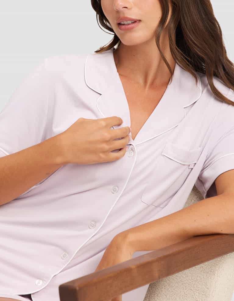 A woman wearing the HIDE Women's Bamboo Stretch-Knit Short Sleeve Pajama Top with white piping sits on a chair, one arm on the wooden armrest and her other hand lightly touching the collar. |Color:Lilac