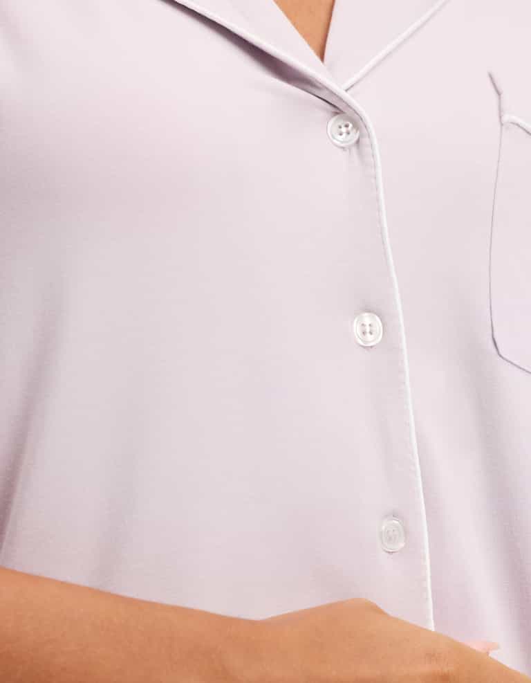 Close-up of HIDE Women's Bamboo Stretch-Knit Short Sleeve Pajama Top in light lavender, featuring white piping, buttons, and a partial chest pocket; part of a person's arm and hand are visible at the bottom. |Color:Lilac