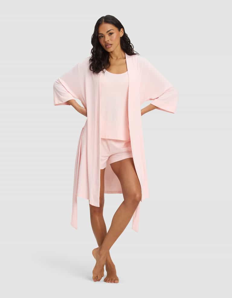 A woman stands barefoot against a gray background, wearing the Cozy Earth Women's Bamboo Stretch-Knit Kimono Robe—a light pink, open-front robe with wide sleeves—over a matching tank and shorts loungewear set.|Color:Blossom