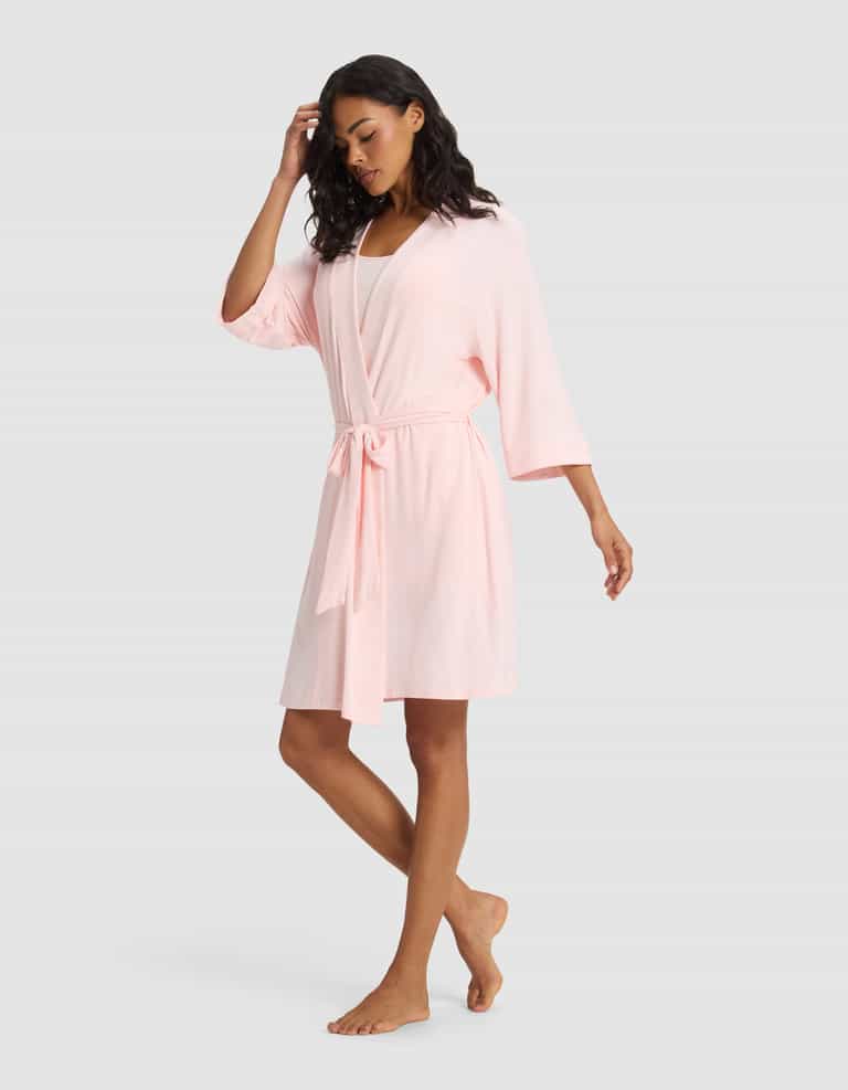 A woman stands barefoot against a plain background, wearing the Cozy Earth Women's Bamboo Stretch-Knit Kimono Robe in light pink, featuring wide sleeves and a waist tie. One hand touches her hair while the other rests by her side.|Color:Blossom