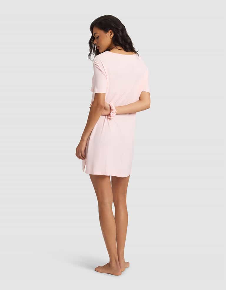 A woman with long dark hair stands barefoot, facing away in a light pink Cozy Earth Women's Bamboo Stretch-Knit Sleep Dress, with one hand behind her back against a plain grey background. |Color:Blossom