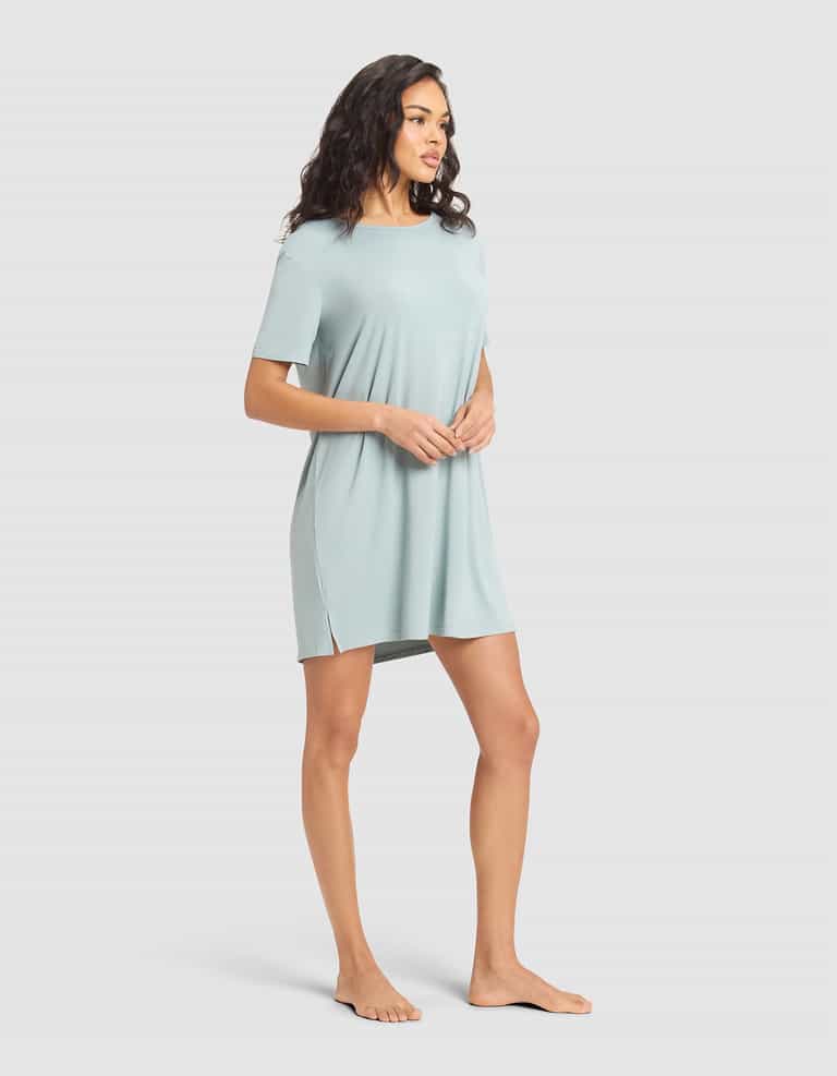A woman stands barefoot against a plain background, wearing the Cozy Earth Women's Bamboo Stretch-Knit Sleep Dress—a relaxed-fit, short-sleeved, light blue dress with side slits—looking to her left with hands clasped in front. |Color:Brook