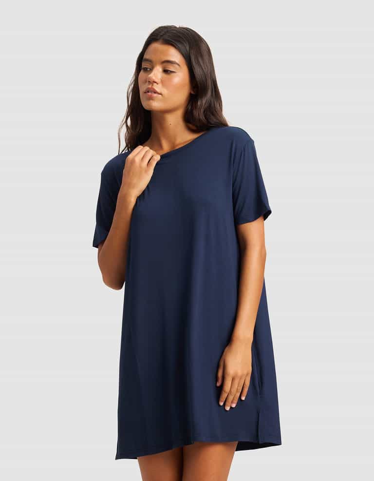 A woman stands against a light gray background, wearing Cozy Earth's Women's Bamboo Stretch-Knit Sleep Dress in navy blue. With long dark hair, she gazes to the side and lightly touches her collar.|Color:Navy