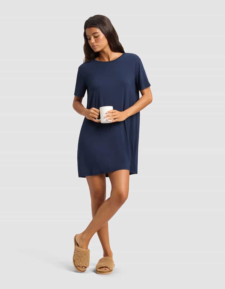A woman in a Cozy Earth Women's Bamboo Stretch-Knit Sleep Dress stands holding a white mug, wearing tan fuzzy slippers. She has long dark hair and looks relaxed against a plain light gray background.|Color:Navy