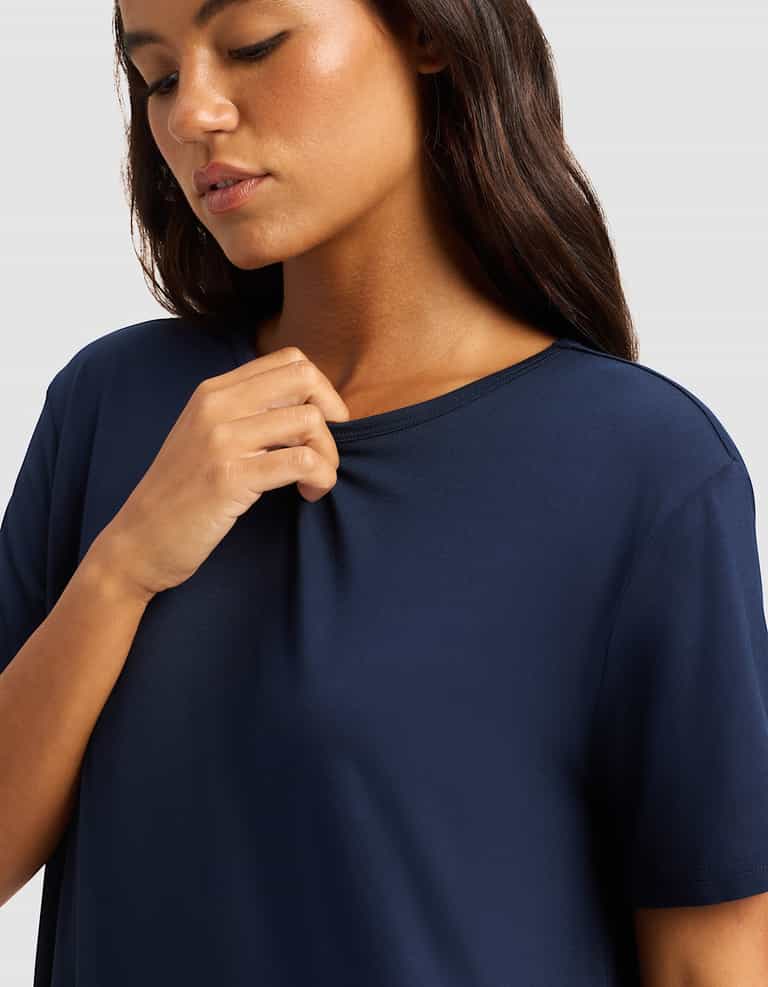 A woman with long brown hair wears the Cozy Earth Women's Bamboo Stretch-Knit Sleep Dress in navy blue, gently holding the collar with her right hand and looking downward calmly against a plain light gray background.|Color:Navy