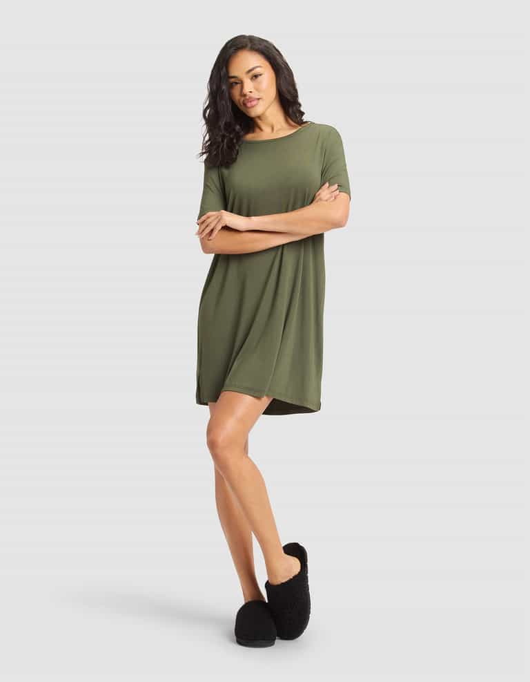 A woman with long, wavy hair wears the Cozy Earth Women's Bamboo Stretch-Knit Sleep Dress in olive green and black fuzzy slippers, standing against a plain light gray background with one leg slightly crossed. |Color:Olive