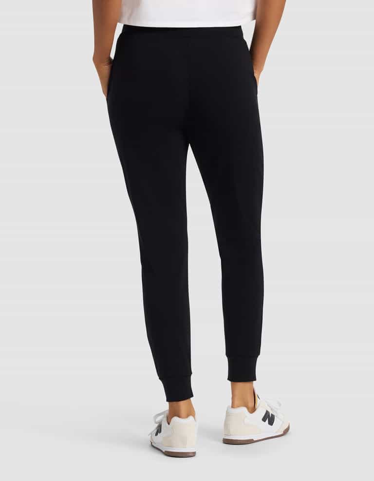 A woman is shown from the back wearing Cozy Earth’s Women's Brushed Bamboo Jogger Pant in black, paired with white sneakers featuring a black "N" logo and a partially visible white shirt, set against a plain light background.|Color:Black