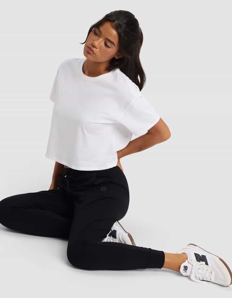 A person wearing a white cropped T-shirt, Cozy Earth Women's Brushed Bamboo Jogger Pant in black, and white New Balance sneakers sits on the floor with one knee up, looking down with a neutral expression against a plain background.|Color:Black