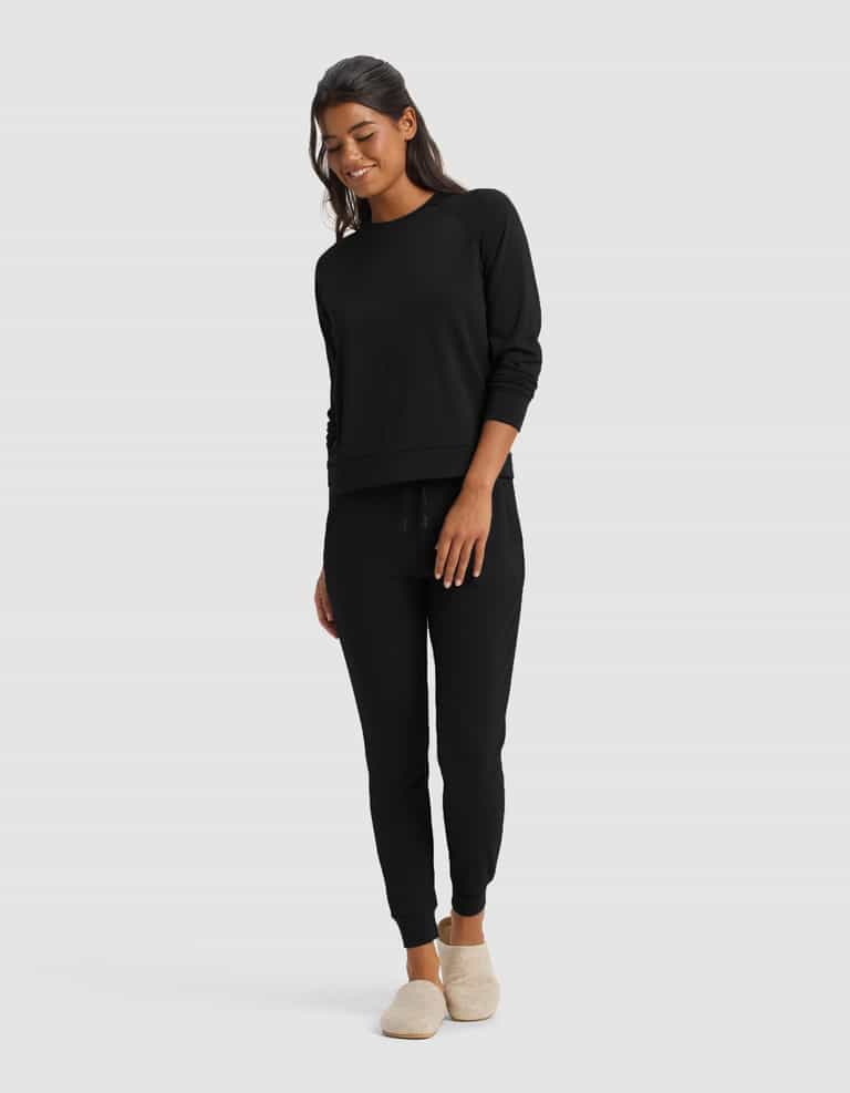 A woman smiles, wearing the Cozy Earth Women's Brushed Bamboo Jogger Set in black with jogger pants and beige slippers, standing against a plain light gray background.|Color:Black