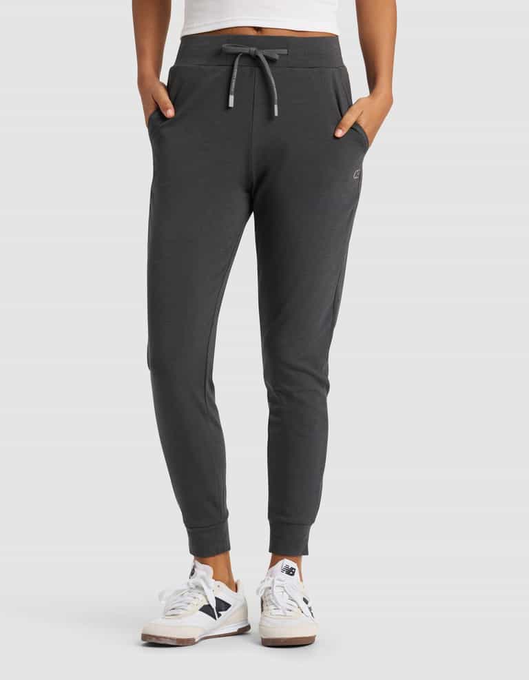 A woman wears Cozy Earth's Women's Brushed Bamboo Jogger Pant in dark gray with white sneakers featuring black accents, standing hands in pockets against a plain light gray background.|Color:Charcoal