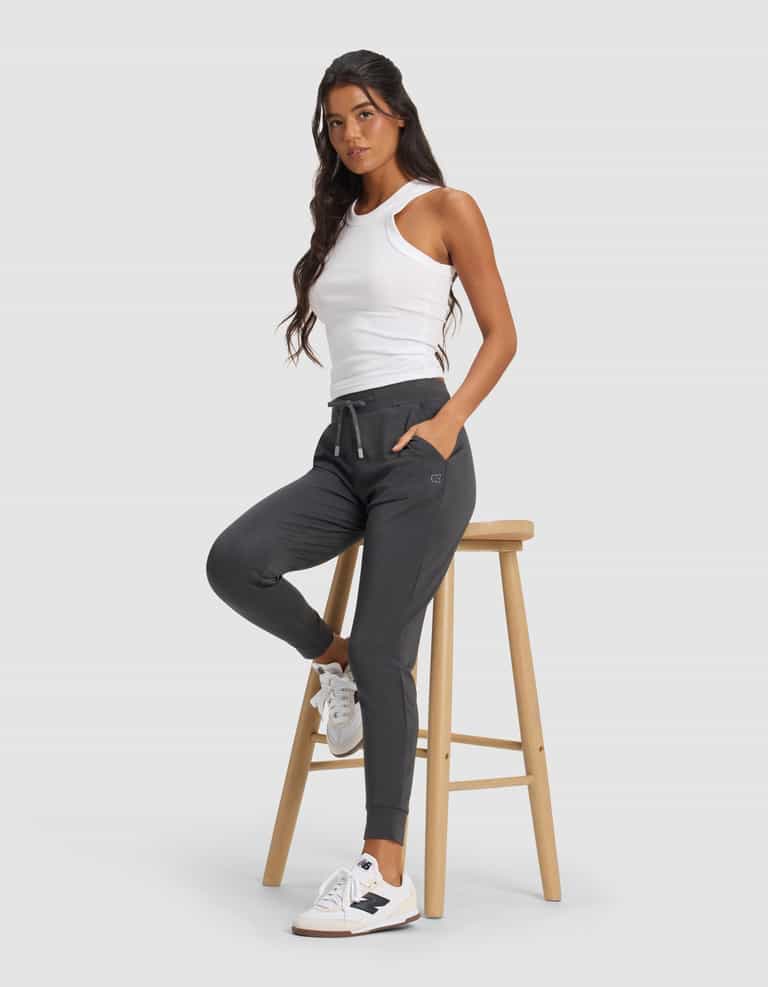 A woman wearing Cozy Earth's Women's Brushed Bamboo Jogger Pant in dark gray poses with one leg on a wooden stool, hands in pockets, and white sneakers, set against a plain light gray background.|Color:Charcoal