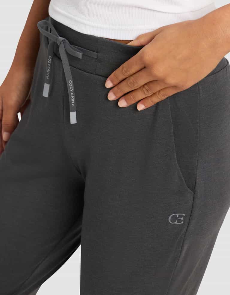 A person wearing the Cozy Earth Women's Brushed Bamboo Jogger Pant in dark gray, featuring side pockets and a small "CE" logo by the left pocket, pairs it with a white top and rests a hand on their hip.|Color:Charcoal