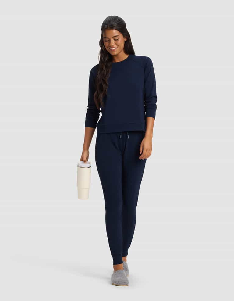 A woman with long dark hair smiles against a plain light background, wearing Cozy Earth's Women's Brushed Bamboo Jogger Pant in navy blue, paired with a matching sweatshirt, gray slippers, and holding a cream-colored tumbler.|Color:Navy